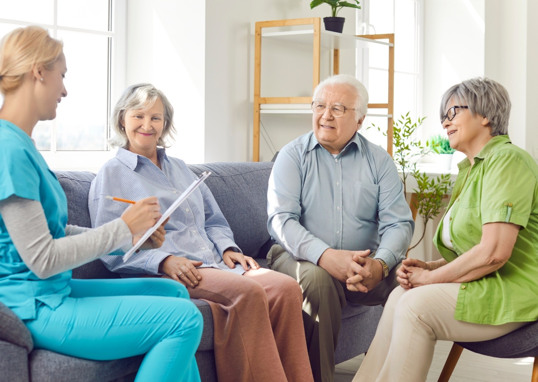 Eine Ärztin in blauer Uniform spricht mit drei älteren Personen auf einem Sofa. Das Gespräch findet in einem hellen Raum statt, der freundlich und modern eingerichtet ist.